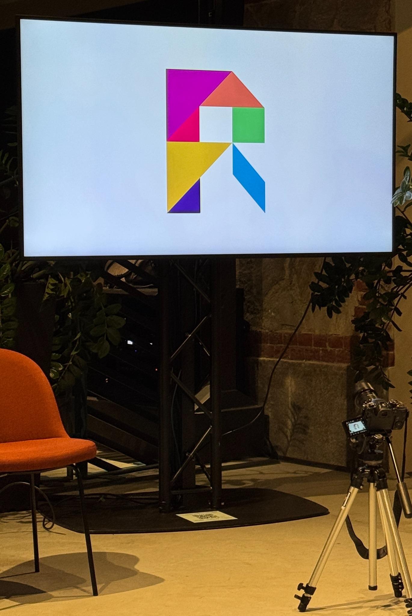 The colourful Rcalls geometric R mark displayed on a screen at the meetup, with a camera on a tripod in the foreground.